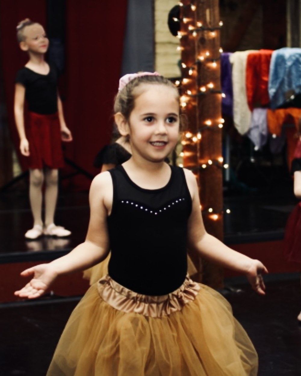 A young ballerina in a gold tutu, arms open on stage, with her class behind her