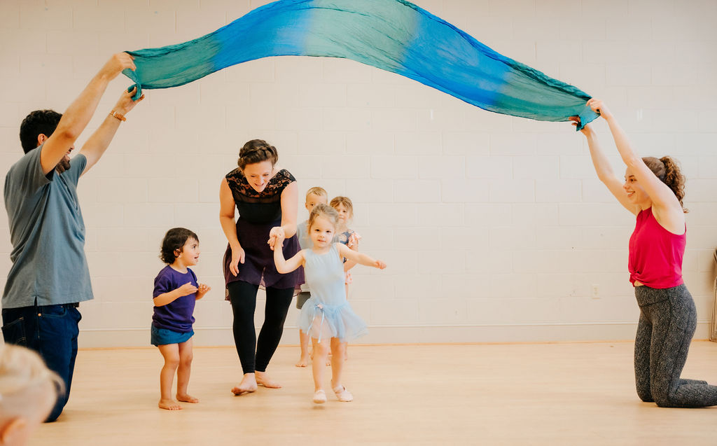 A class of young dancers moving together under a flowing silk with their instructor